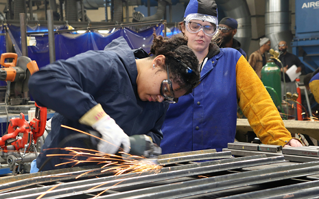 Participant uses a grinder on metal while an instructor observes during a hands-on industrial training session.
